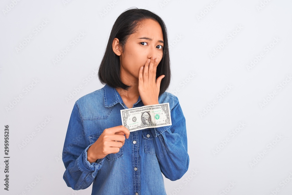 Young beautiful chinese woman holding one dollar standing over isolated white background cover mouth with hand shocked with shame for mistake, expression of fear, scared in silence, secret concept Young beautiful chinese woman holding one dollar standing over isolated white background cover mouth with hand shocked with shame for mistake, expression of fear, scared in silence, secret concept