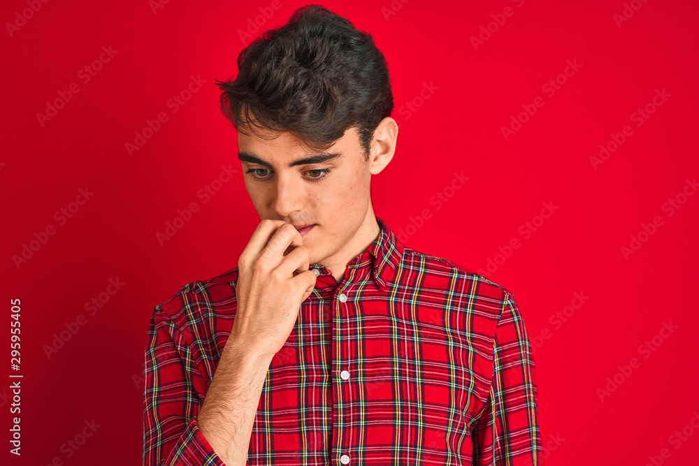 Teenager boy wearing red shirt standing over isolated background looking stressed and nervous with hands on mouth biting nails. Anxiety problem.