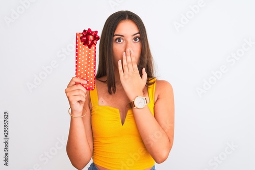 Young beautiful girl holding birthday gift standing over isolated white background cover mouth with hand shocked with shame for mistake, expression of fear, scared in silence, secret concept