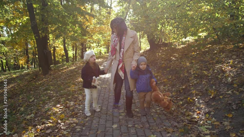 Cheerful mother communicating with children while walking hand in hand along sunlit park covered with yellow autumn leaves. Happy mom with special needs girl and toddler boy enjoying freetime outdoor Cheerful mother communicating with children while walking hand in hand along sunlit park covered with yellow autumn leaves. Happy mom with special needs girl and toddler boy enjoying freetime outdoor