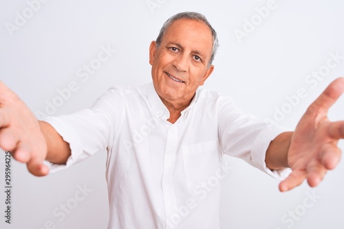 Senior grey-haired man wearing elegant shirt standing over isolated white background looking at the camera smiling with open arms for hug. Cheerful expression embracing happiness.