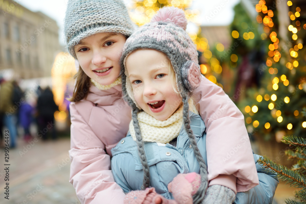 Fototapeta premium Two adorable sisters having a good time together on traditional Christmas fair in Riga, Latvia. Children enjoying sweets, candies and gingerbread on Xmas market.