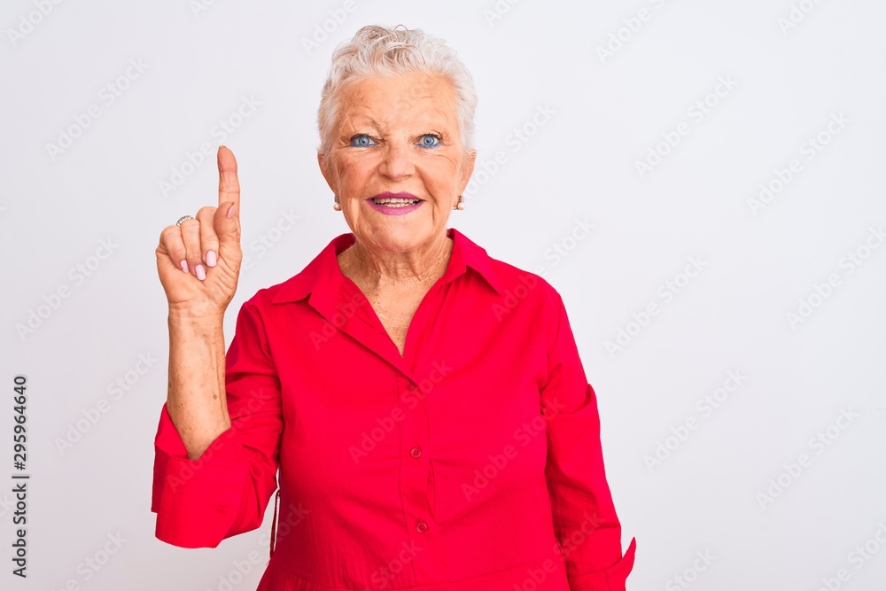 Senior grey-haired woman wearing red casual shirt standing over isolated white background showing and pointing up with finger number one while smiling confident and happy. Senior grey-haired woman wearing red casual shirt standing over isolated white background showing and pointing up with finger number one while smiling confident and happy.