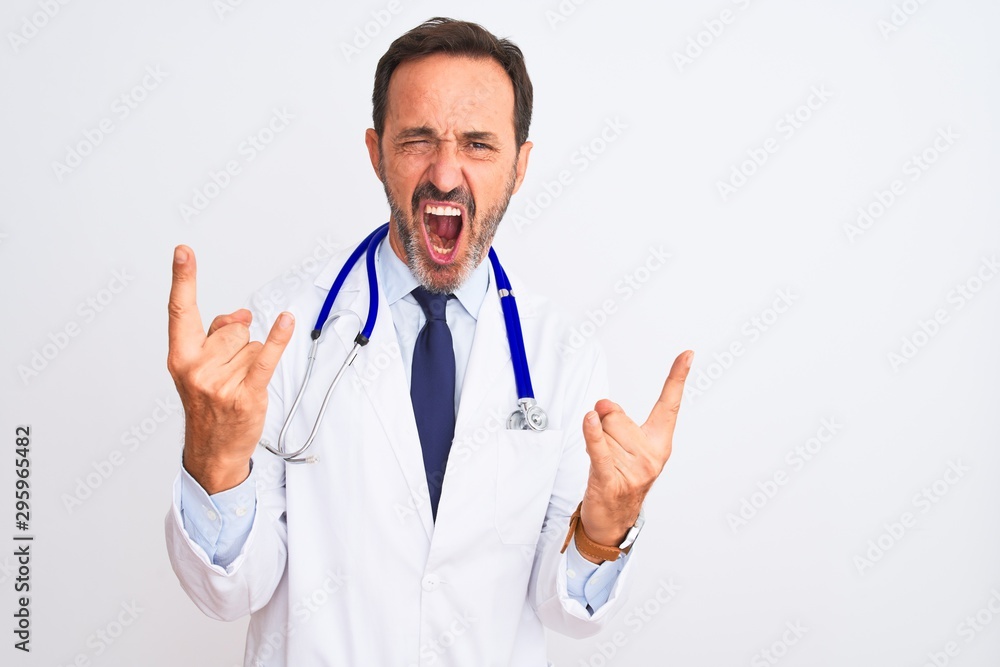 Middle age doctor man wearing coat and stethoscope standing over isolated white background shouting with crazy expression doing rock symbol with hands up. Music star. Heavy concept. Middle age doctor man wearing coat and stethoscope standing over isolated white background shouting with crazy expression doing rock symbol with hands up. Music star. Heavy concept.