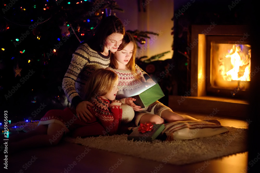 Happy young mother and her two small daughters opening a magical Christmas gift by a fireplace in a cozy dark living room on Christmas eve.