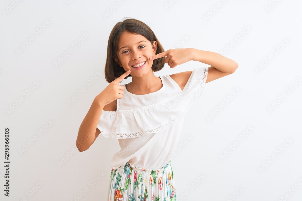 Young beautiful child girl wearing casual dress standing over isolated white background smiling cheerful showing and pointing with fingers teeth and mouth. Dental health concept. Young beautiful child girl wearing casual dress standing over isolated white background smiling cheerful showing and pointing with fingers teeth and mouth. Dental health concept.