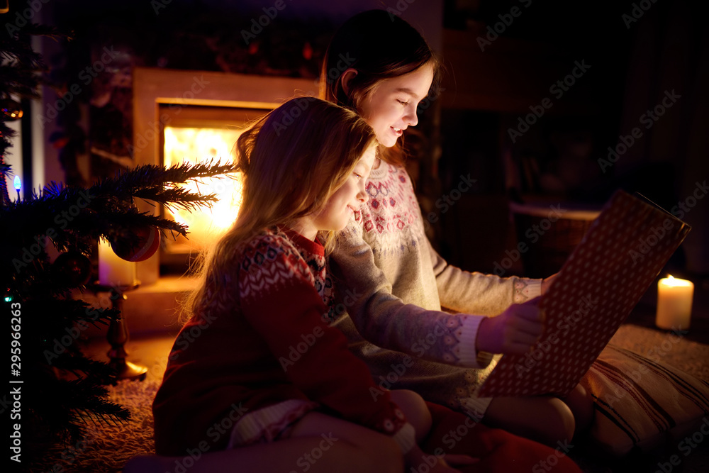 Happy young sisters reading a story book together by a fireplace in a cozy dark living room on Christmas eve. Celebrating Xmas at home. Happy young sisters reading a story book together by a fireplace in a cozy dark living room on Christmas eve. Celebrating Xmas at home.