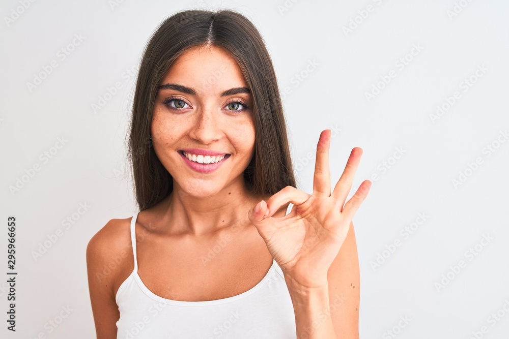 Young beautiful woman wearing casual t-shirt standing over isolated white background doing ok sign with fingers, excellent symbol Young beautiful woman wearing casual t-shirt standing over isolated white background doing ok sign with fingers, excellent symbol