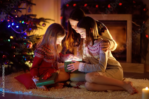 Happy young mother and her two small daughters opening a magical Christmas gift by a fireplace in a cozy dark living room on Christmas eve.