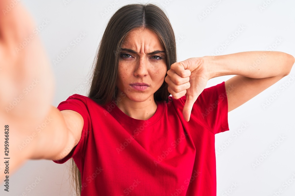 Beautiful woman wearing red t-shirt make selfie by camera over isolated white background with angry face, negative sign showing dislike with thumbs down, rejection concept Beautiful woman wearing red t-shirt make selfie by camera over isolated white background with angry face, negative sign showing dislike with thumbs down, rejection concept