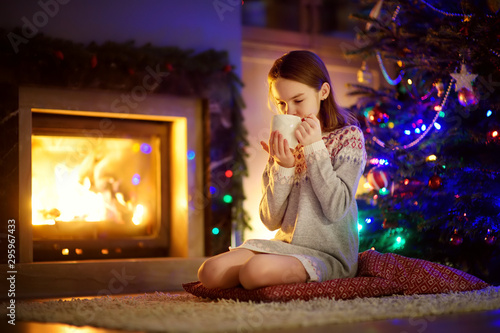 Happy young girl having a cup of hot chocolate by a fireplace in a cozy dark living room on Christmas eve. Celebrating Xmas at home.
