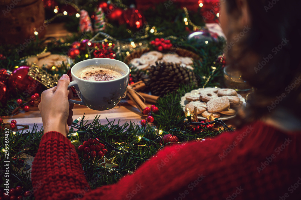 Young woman holding a cup of hot chocolate at Christmas and in the background a table with holiday season ingredients like orange, cookies, panettone, cinnamon and pine Young woman holding a cup of hot chocolate at Christmas and in the background a table with holiday season ingredients like orange, cookies, panettone, cinnamon and pine