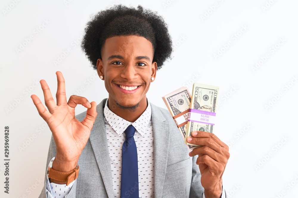 Young african american businessman holding dollars standing over isolated white background doing ok sign with fingers, excellent symbol Young african american businessman holding dollars standing over isolated white background doing ok sign with fingers, excellent symbol