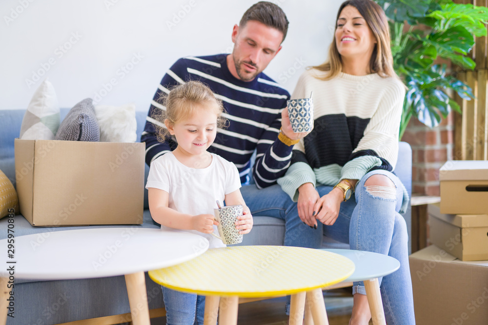 Beautiful family, parents sitting on the sofa drinking coffee looking his kid playing at new home around cardboard boxes Beautiful family, parents sitting on the sofa drinking coffee looking his kid playing at new home around cardboard boxes