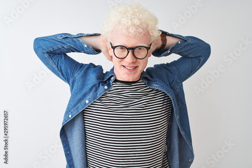 Young albino blond man wearing denim shirt and glasses over isolated white background relaxing and stretching, arms and hands behind head and neck smiling happy