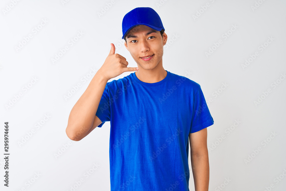 Chinese deliveryman wearing blue t-shirt and cap standing over isolated white background smiling doing phone gesture with hand and fingers like talking on the telephone. Communicating concepts.