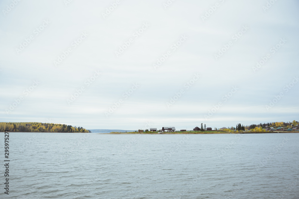 autumn landscape with surface lake water, country village, yellow deciduous and green coniferous forest, and cloudy sky, horizontal stock photo image