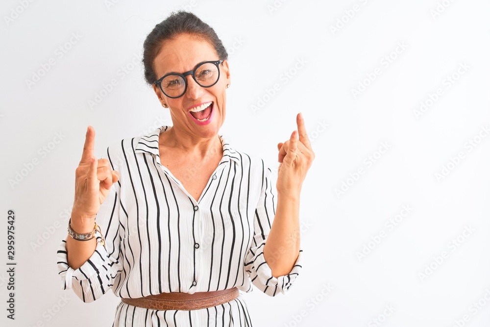 Middle age businesswoman wearing striped dress and glasses over isolated white background shouting with crazy expression doing rock symbol with hands up. Music star. Heavy music concept. Middle age businesswoman wearing striped dress and glasses over isolated white background shouting with crazy expression doing rock symbol with hands up. Music star. Heavy music concept.