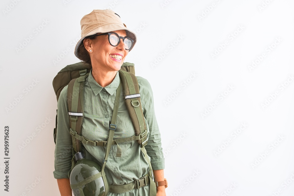Middle age hiker woman wearing backpack canteen hat glasses over isolated white background looking away to side with smile on face, natural expression. Laughing confident. Middle age hiker woman wearing backpack canteen hat glasses over isolated white background looking away to side with smile on face, natural expression. Laughing confident.