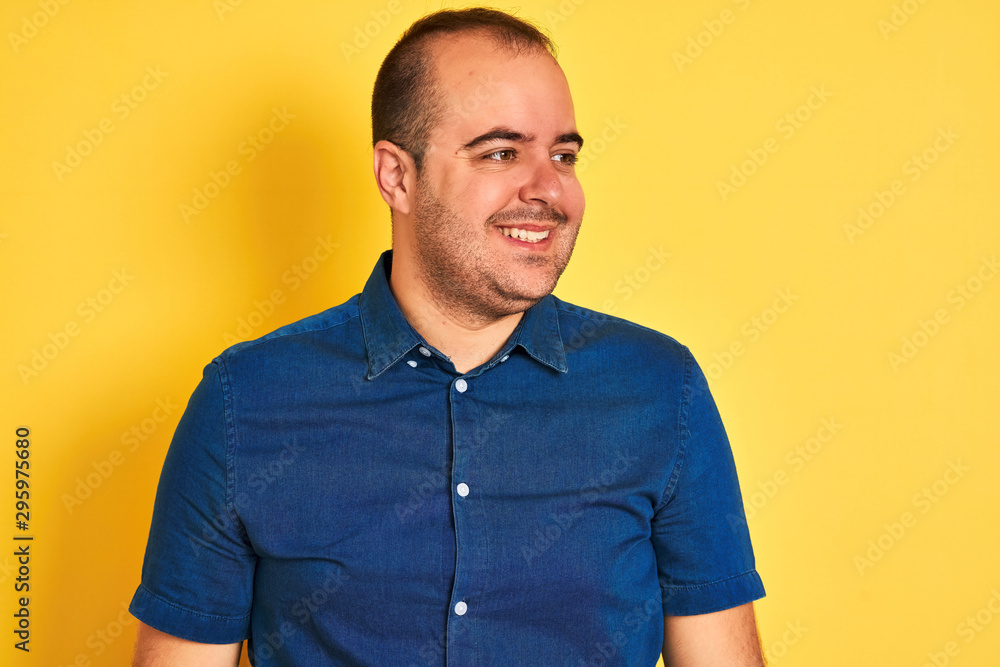 Young man wearing denim casual shirt standing over isolated yellow background looking away to side with smile on face, natural expression. Laughing confident. Young man wearing denim casual shirt standing over isolated yellow background looking away to side with smile on face, natural expression. Laughing confident.