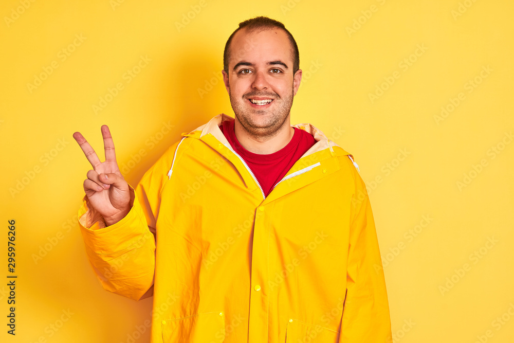 Naklejka premium Young man wearing rain coat standing over isolated yellow background smiling with happy face winking at the camera doing victory sign. Number two.