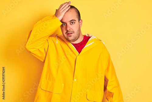 Young man wearing rain coat standing over isolated yellow background surprised with hand on head for mistake  remember error. Forgot  bad memory concept.