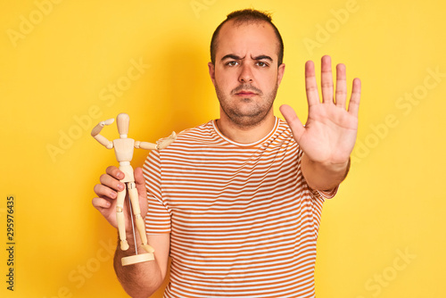 Young man holding figure of art dummy standing over isolated yellow background with open hand doing stop sign with serious and confident expression, defense gesture