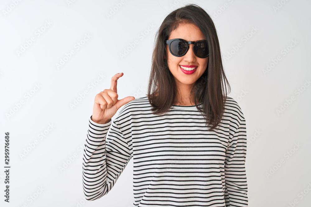 Chinese woman wearing striped t-shirt and sunglasses standing over isolated white background smiling and confident gesturing with hand doing small size sign with fingers looking and the camera Chinese woman wearing striped t-shirt and sunglasses standing over isolated white background smiling and confident gesturing with hand doing small size sign with fingers looking and the camera