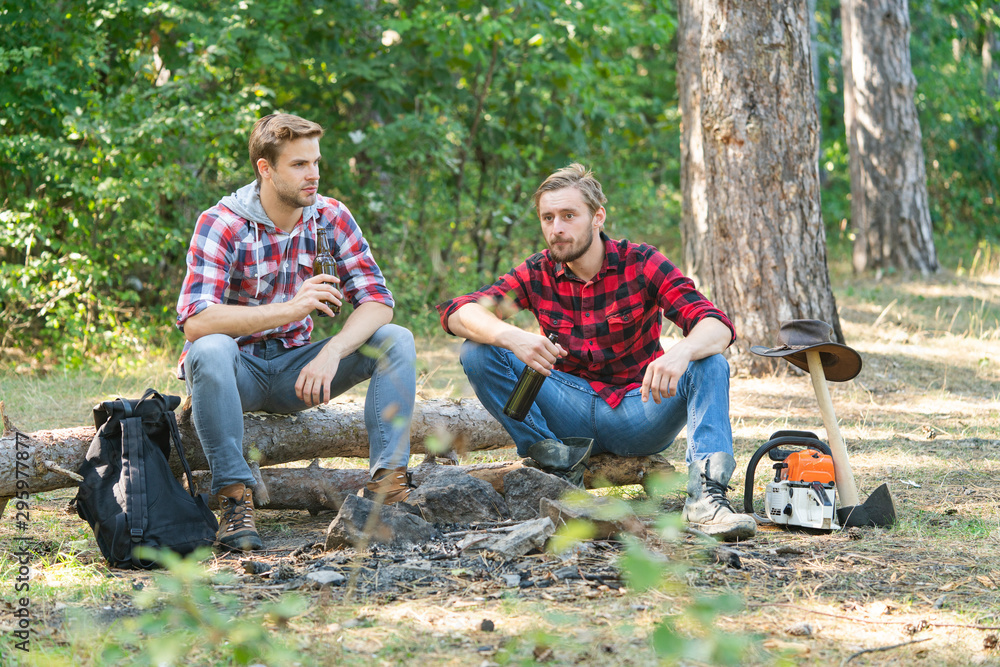 Camp adventure and travel concept. Tourists relaxing. Group of friends enjoying picnic in the forest and drinking beer. Two friends lumberjack worker sitting in the forest and drinking beer. Camp adventure and travel concept. Tourists relaxing. Group of friends enjoying picnic in the forest and drinking beer. Two friends lumberjack worker sitting in the forest and drinking beer.