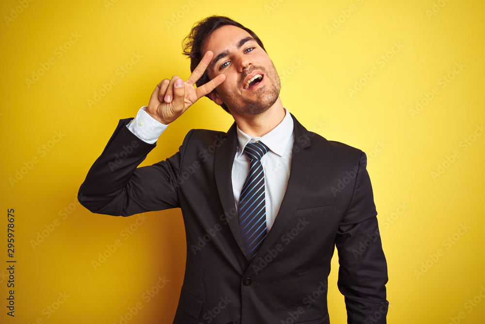 Young handsome businessman wearing suit and tie standing over isolated yellow background Doing peace symbol with fingers over face, smiling cheerful showing victory Young handsome businessman wearing suit and tie standing over isolated yellow background Doing peace symbol with fingers over face, smiling cheerful showing victory