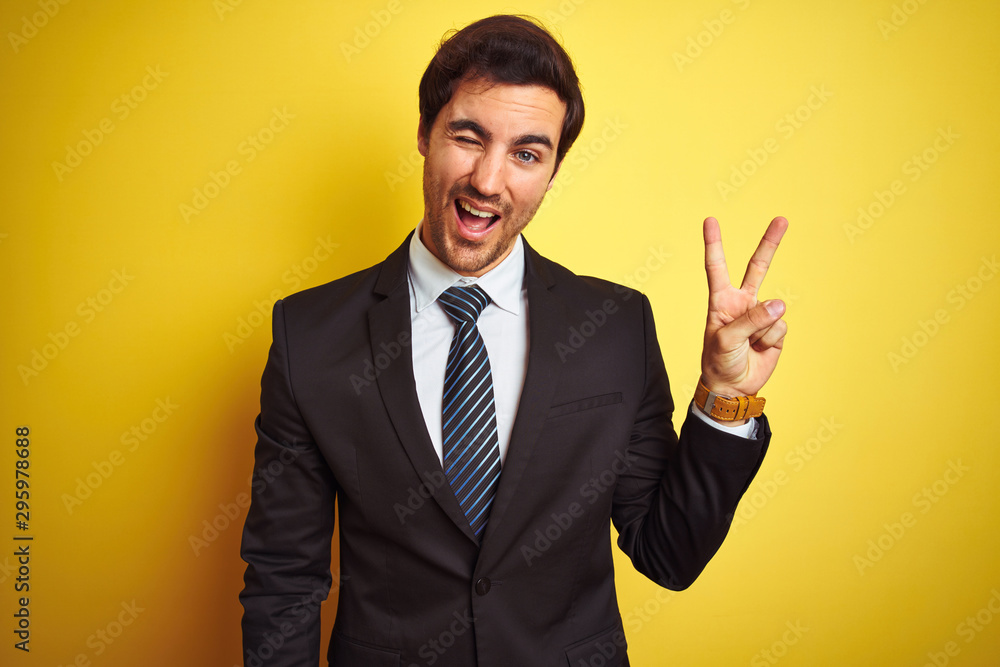 Young handsome businessman wearing suit and tie standing over isolated yellow background smiling with happy face winking at the camera doing victory sign with fingers. Number two. Young handsome businessman wearing suit and tie standing over isolated yellow background smiling with happy face winking at the camera doing victory sign with fingers. Number two.