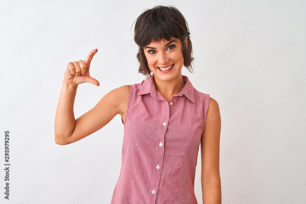 Young beautiful woman wearing red summer shirt standing over isolated white background smiling and confident gesturing with hand doing small size sign with fingers looking and the camera Young beautiful woman wearing red summer shirt standing over isolated white background smiling and confident gesturing with hand doing small size sign with fingers looking and the camera