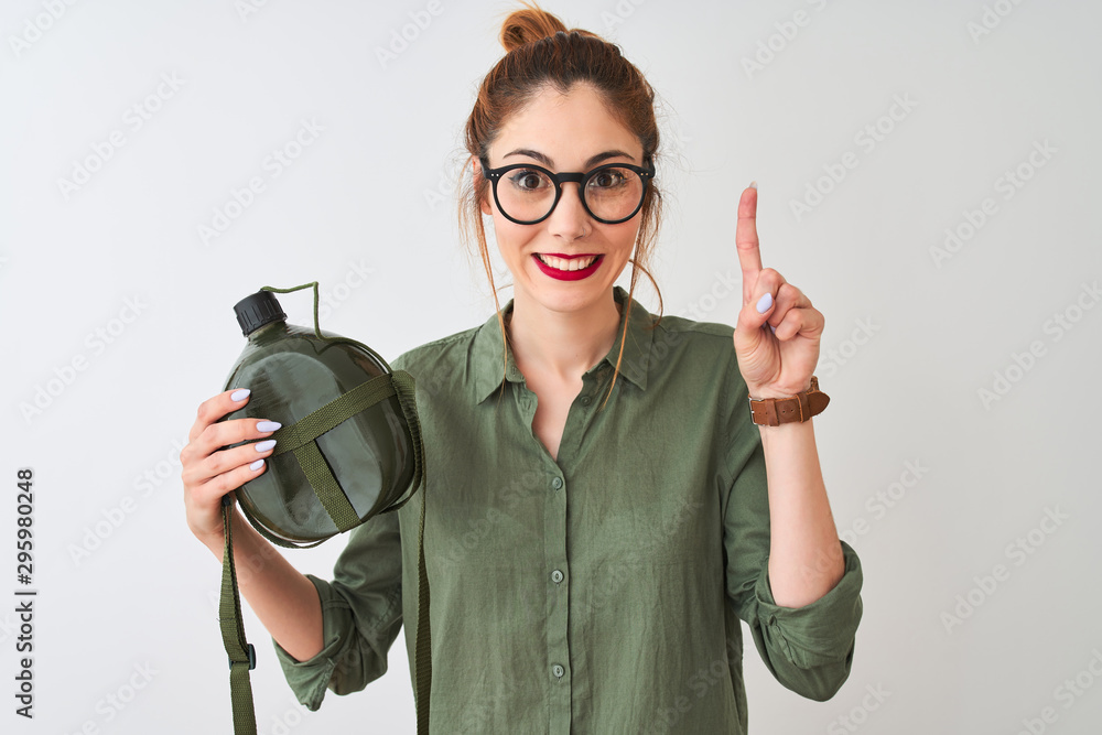 Beautiful redhead woman wearing glasses holding canteen over isolated white background surprised with an idea or question pointing finger with happy face, number one Beautiful redhead woman wearing glasses holding canteen over isolated white background surprised with an idea or question pointing finger with happy face, number one