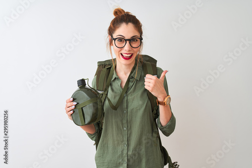 Redhead hiker woman wearing backpack holding canteen over isolated white background pointing and showing with thumb up to the side with happy face smiling