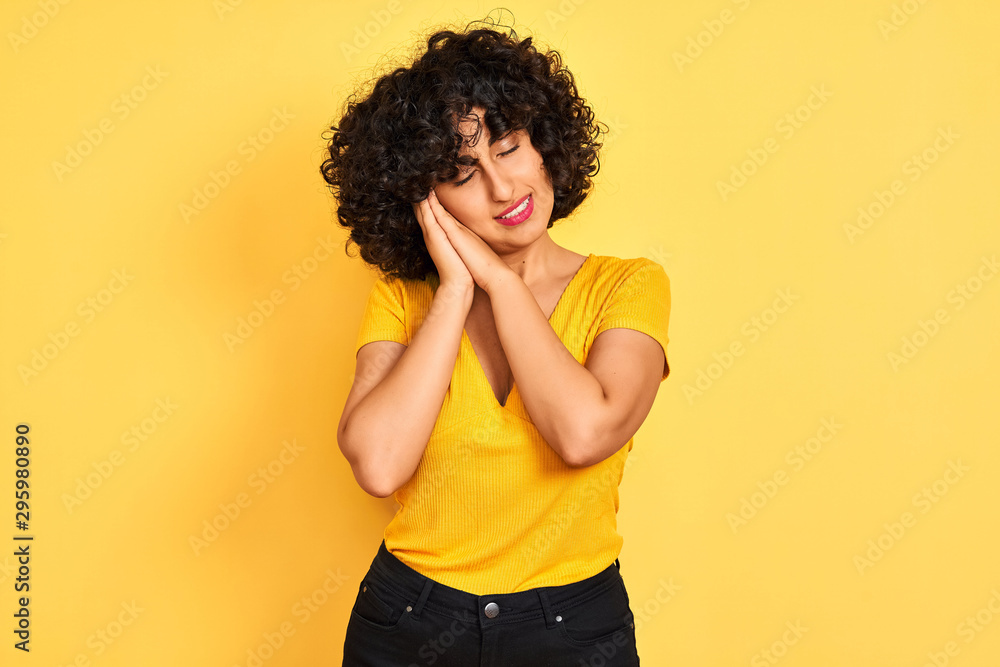 Young arab woman with curly hair wearing t-shirt standing over isolated yellow background sleeping tired dreaming and posing with hands together while smiling with closed eyes. Young arab woman with curly hair wearing t-shirt standing over isolated yellow background sleeping tired dreaming and posing with hands together while smiling with closed eyes.