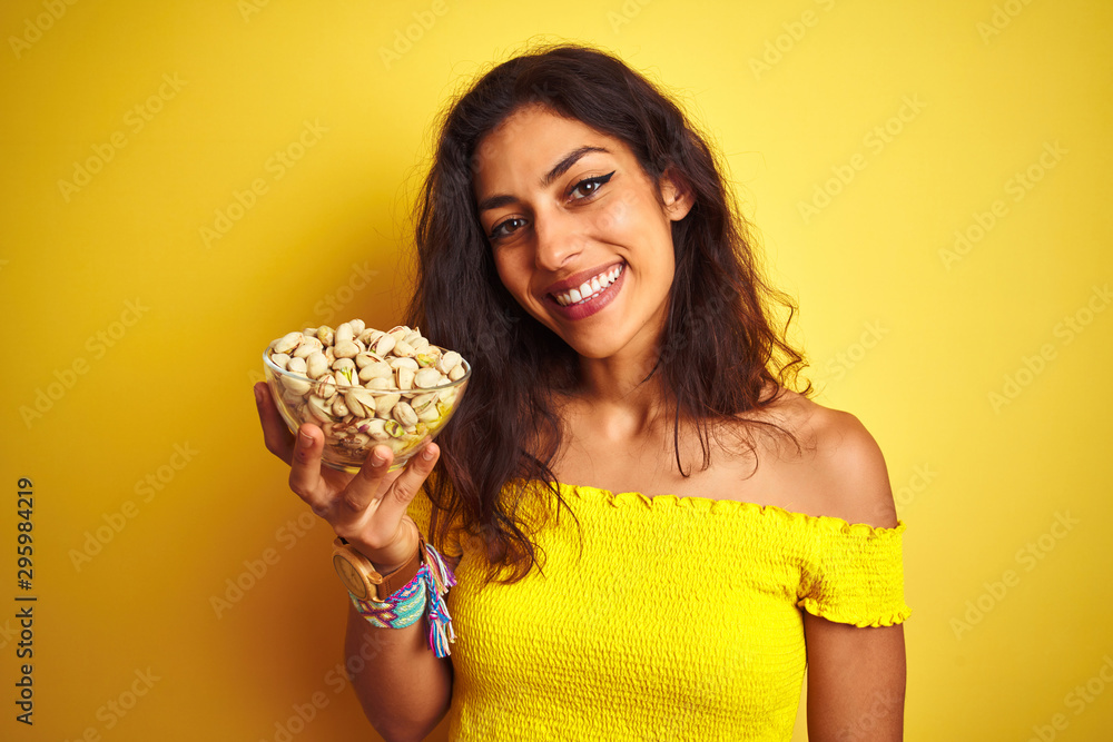 Young beautiful woman holding bowl with pistachios over isolated yellow background with a happy face standing and smiling with a confident smile showing teeth Young beautiful woman holding bowl with pistachios over isolated yellow background with a happy face standing and smiling with a confident smile showing teeth