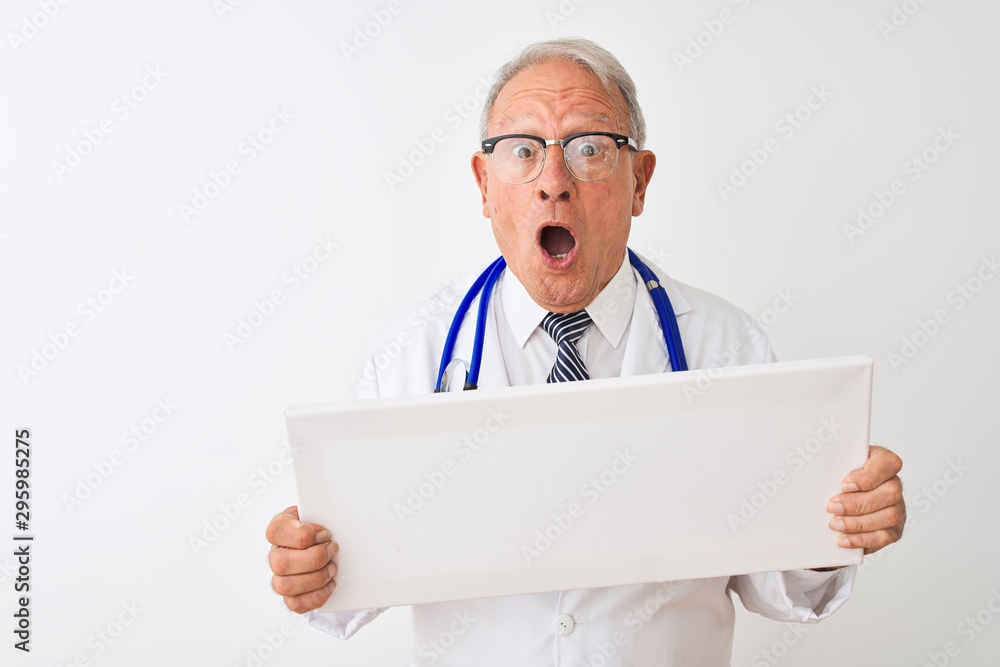 Senior grey-haired doctor man holding banner standing over isolated white background scared in shock with a surprise face, afraid and excited with fear expression Senior grey-haired doctor man holding banner standing over isolated white background scared in shock with a surprise face, afraid and excited with fear expression