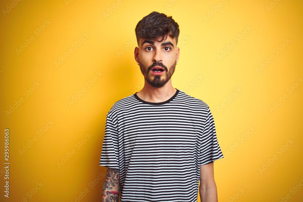 Young man with tattoo wearing striped t-shirt standing over isolated yellow background afraid and shocked with surprise expression, fear and excited face.