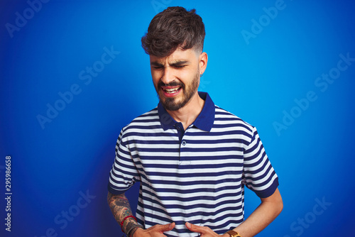 Young man with tattoo wearing striped polo standing over isolated blue background with hand on stomach because indigestion, painful illness feeling unwell. Ache concept.
