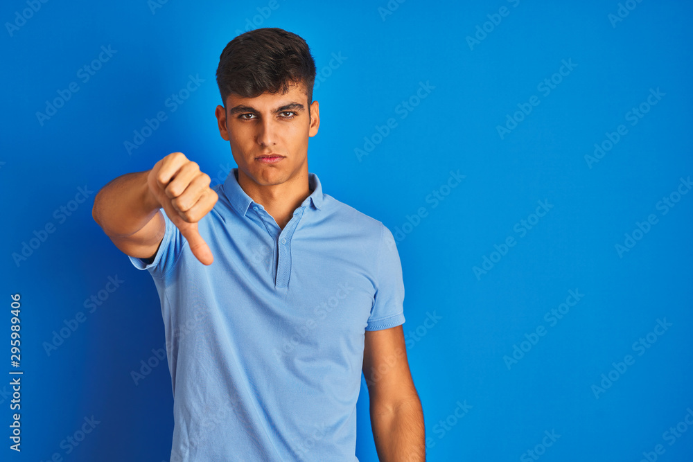 Young indian man wearing casual polo standing over isolated blue background looking unhappy and angry showing rejection and negative with thumbs down gesture. Bad expression. Young indian man wearing casual polo standing over isolated blue background looking unhappy and angry showing rejection and negative with thumbs down gesture. Bad expression.