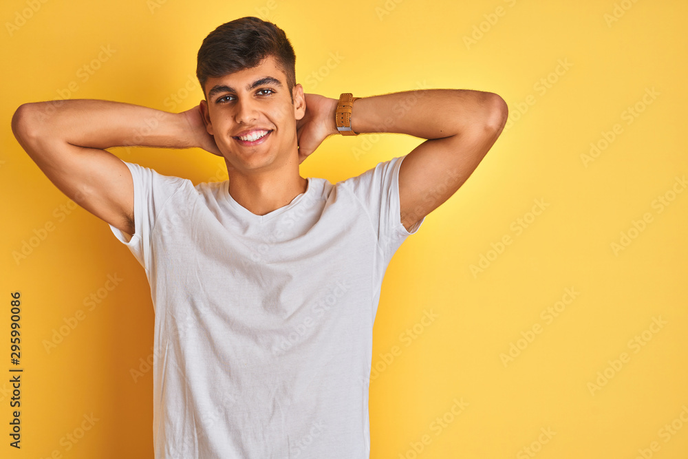 Young indian man wearing white t-shirt standing over isolated yellow background relaxing and stretching, arms and hands behind head and neck smiling happy Young indian man wearing white t-shirt standing over isolated yellow background relaxing and stretching, arms and hands behind head and neck smiling happy