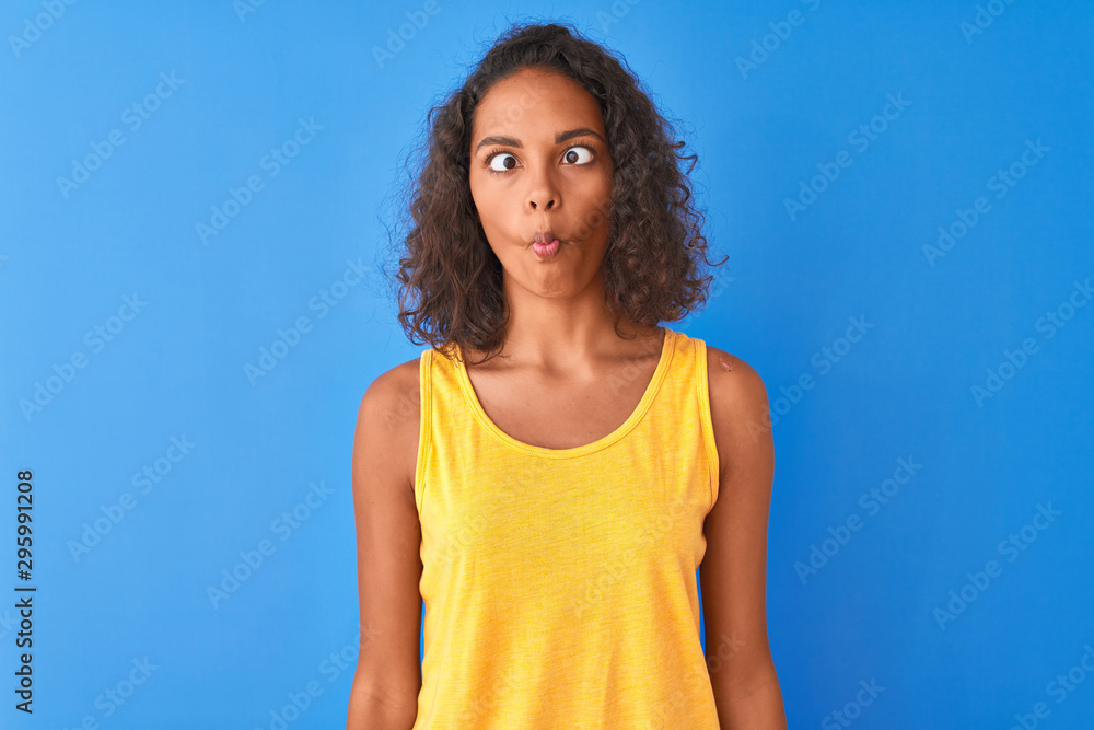 Young brazilian woman wearing yellow t-shirt standing over isolated blue background making fish face with lips, crazy and comical gesture. Funny expression. Young brazilian woman wearing yellow t-shirt standing over isolated blue background making fish face with lips, crazy and comical gesture. Funny expression.
