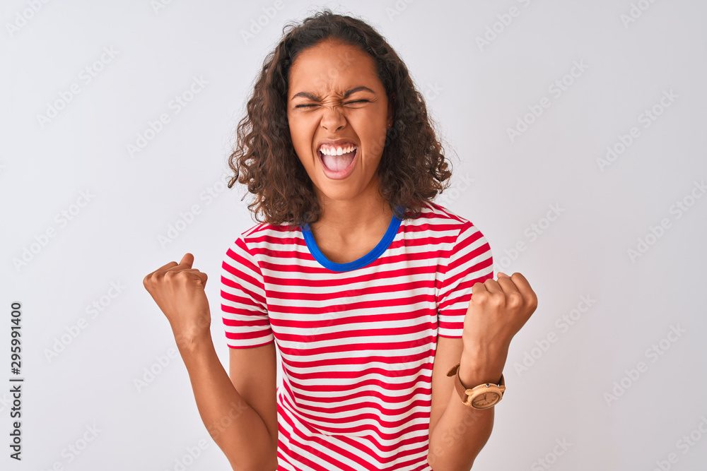 Young brazilian woman wearing red striped t-shirt standing over isolated white background very happy and excited doing winner gesture with arms raised, smiling and screaming for success Young brazilian woman wearing red striped t-shirt standing over isolated white background very happy and excited doing winner gesture with arms raised, smiling and screaming for success