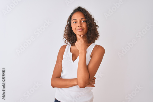 Young brazilian woman wearing casual t-shirt standing over isolated white background looking confident at the camera smiling with crossed arms and hand raised on chin. Thinking positive.
