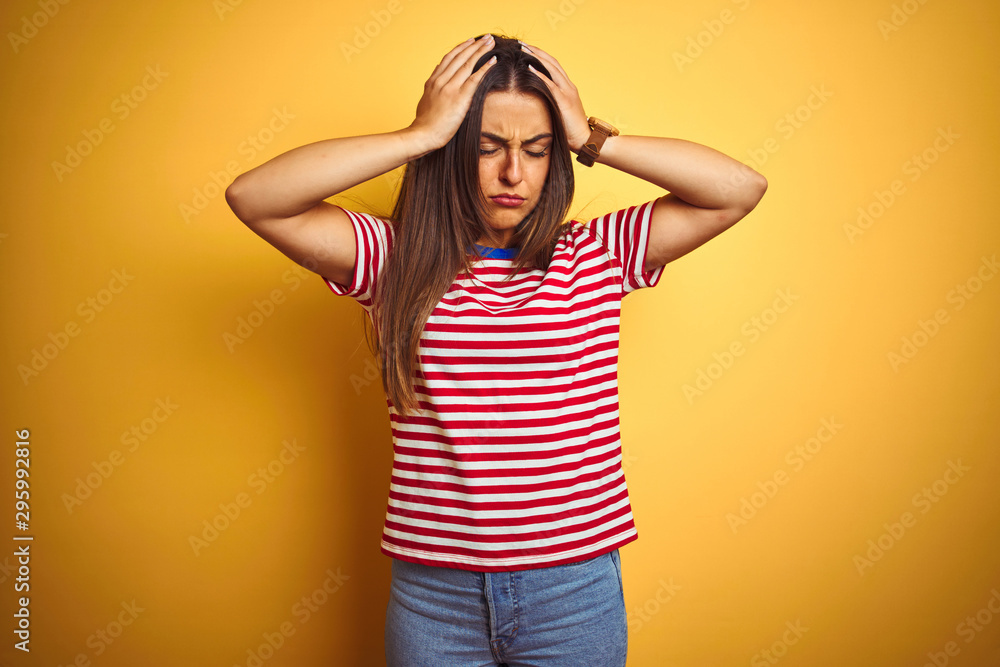 Young beautiful woman wearing striped t-shirt standing over isolated yellow background suffering from headache desperate and stressed because pain and migraine. Hands on head. Young beautiful woman wearing striped t-shirt standing over isolated yellow background suffering from headache desperate and stressed because pain and migraine. Hands on head.