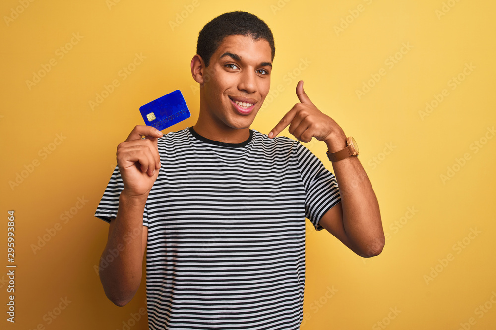 Young handsome arab man holding credit card standing over isolated yellow background with surprise face pointing finger to himself Young handsome arab man holding credit card standing over isolated yellow background with surprise face pointing finger to himself