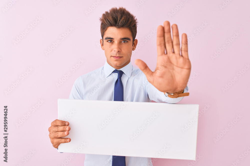 Young handsome businessman holding banner standing over isolated pink background with open hand doing stop sign with serious and confident expression, defense gesture Young handsome businessman holding banner standing over isolated pink background with open hand doing stop sign with serious and confident expression, defense gesture