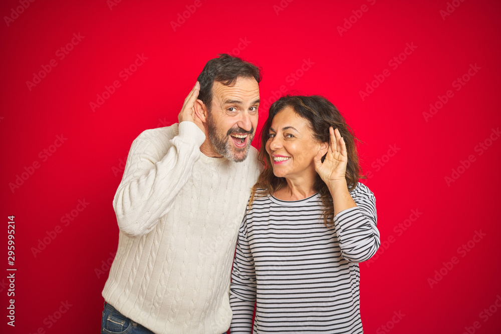 Beautiful middle age couple wearing winter sweater over isolated red background smiling with hand over ear listening an hearing to rumor or gossip. Deafness concept. Beautiful middle age couple wearing winter sweater over isolated red background smiling with hand over ear listening an hearing to rumor or gossip. Deafness concept.