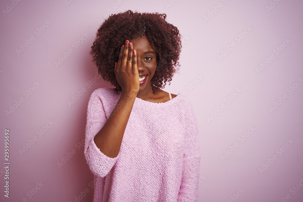 Young african afro woman wearing sweater standing over isolated pink background covering one eye with hand, confident smile on face and surprise emotion. Young african afro woman wearing sweater standing over isolated pink background covering one eye with hand, confident smile on face and surprise emotion.