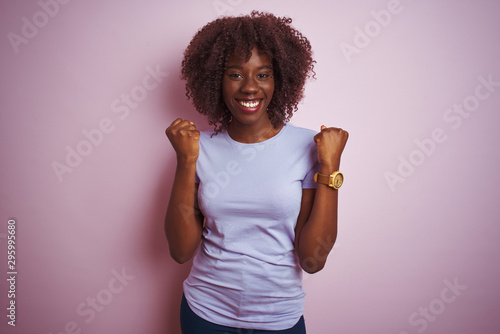 Young african afro woman wearing t-shirt standing over isolated pink background celebrating surprised and amazed for success with arms raised and open eyes. Winner concept.